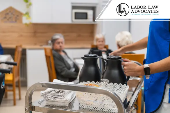A caregiver in a blue uniform serves beverages from a cart to elderly residents seated at a table in a nursing home